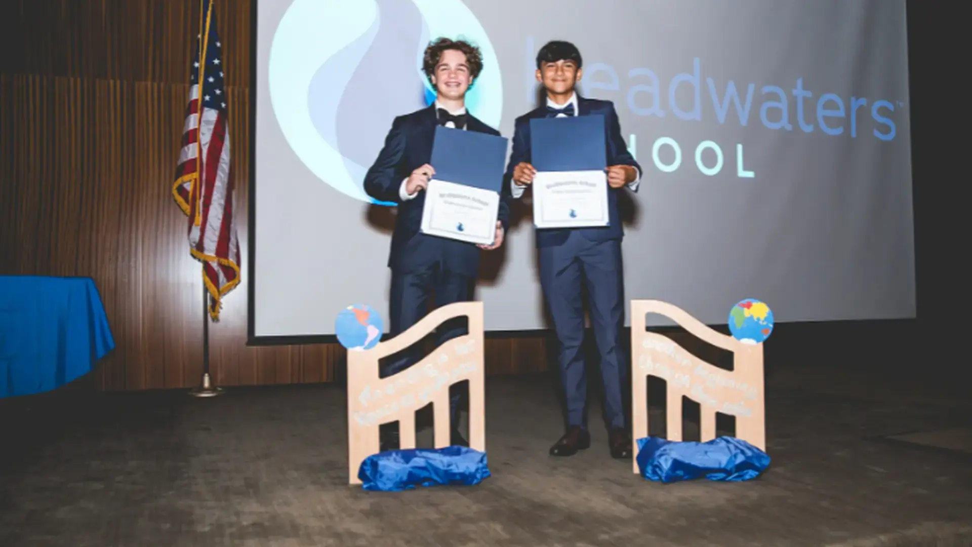 Two graduates standing on stage holding diplomas in front of a Headwaters School backdrop.