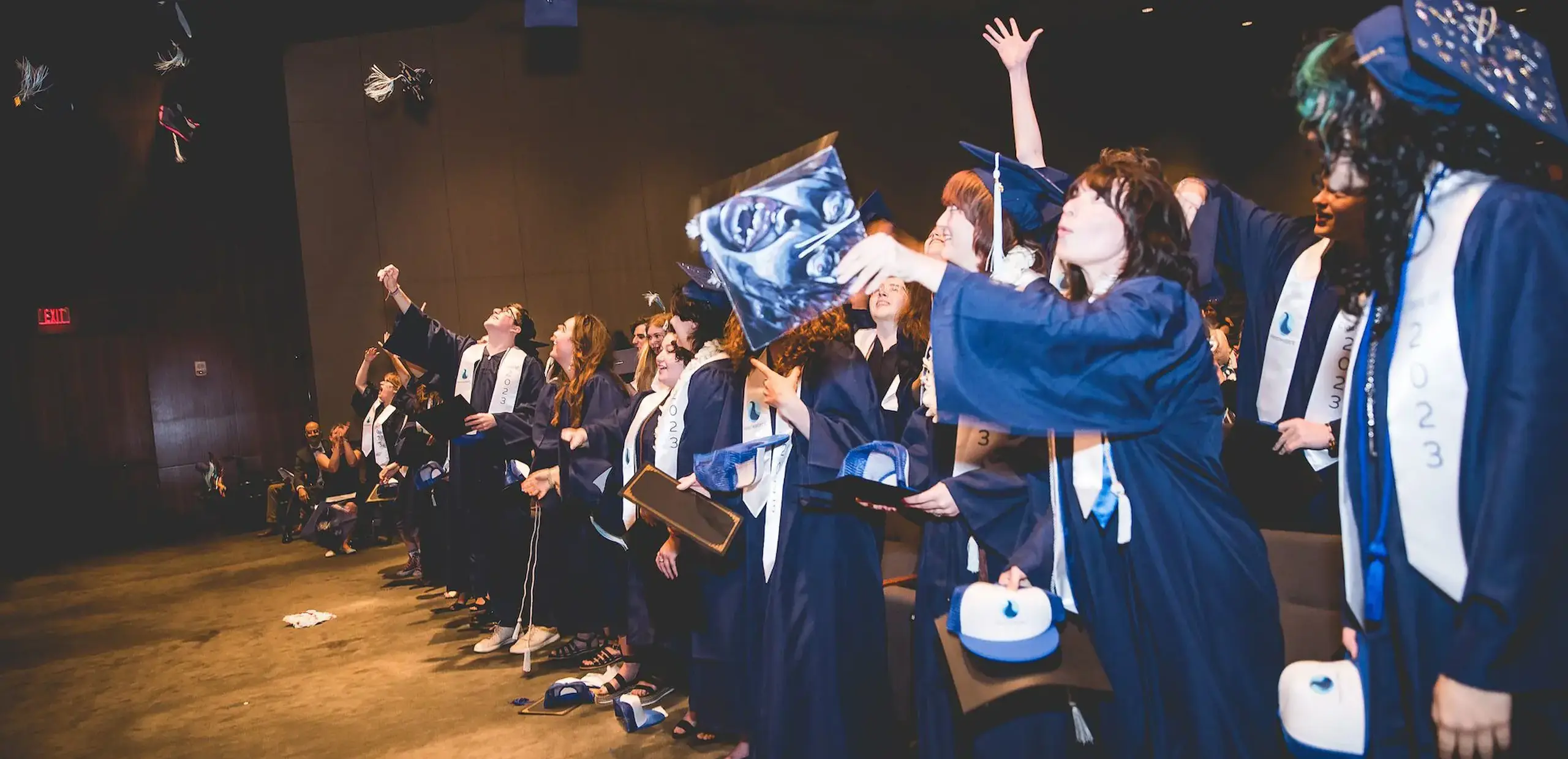 Class of 2023 graduates in navy caps and gowns throwing their caps into the air during commencement.