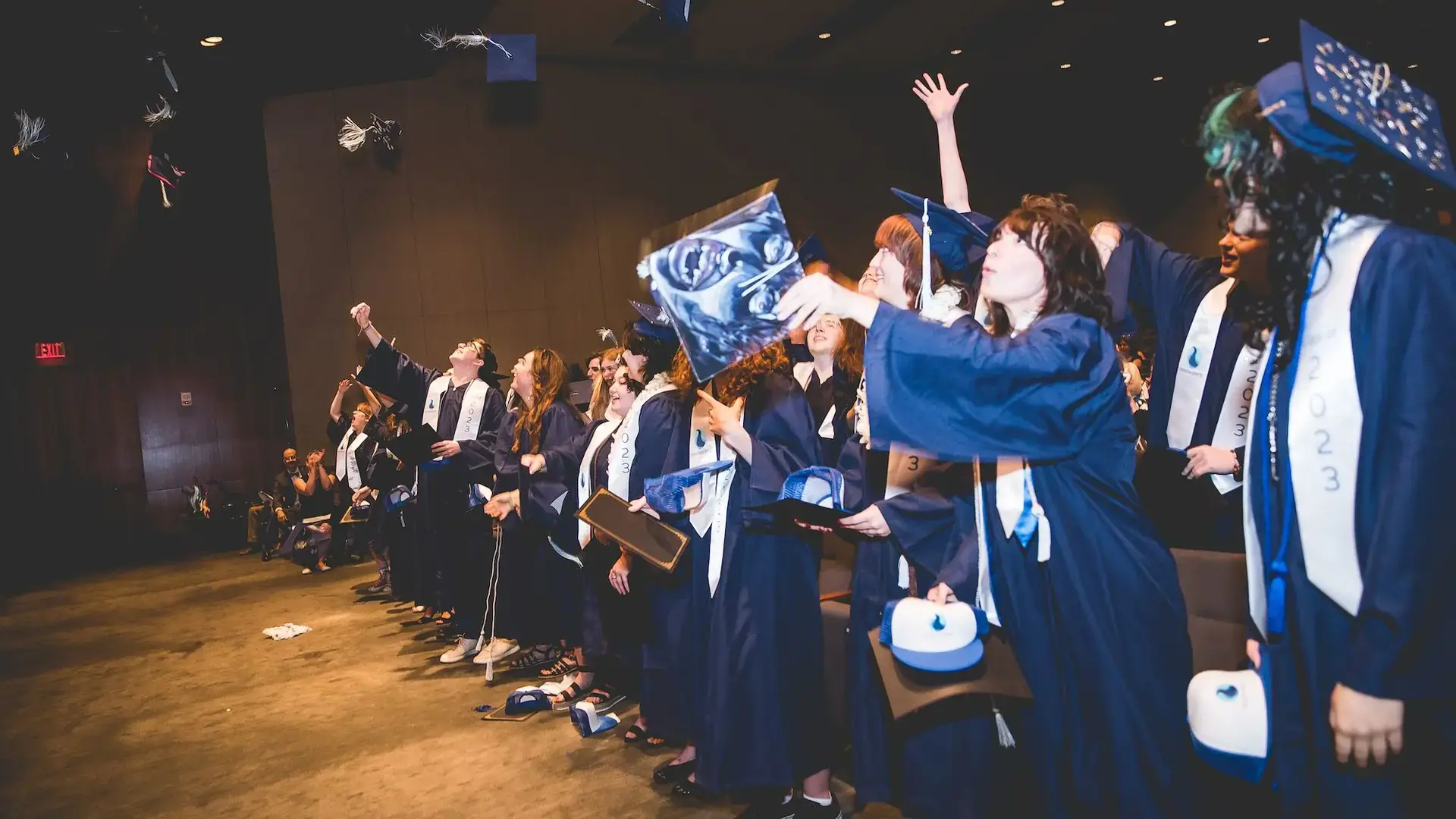 Class of 2023 graduates in navy caps and gowns throwing their caps into the air during commencement.