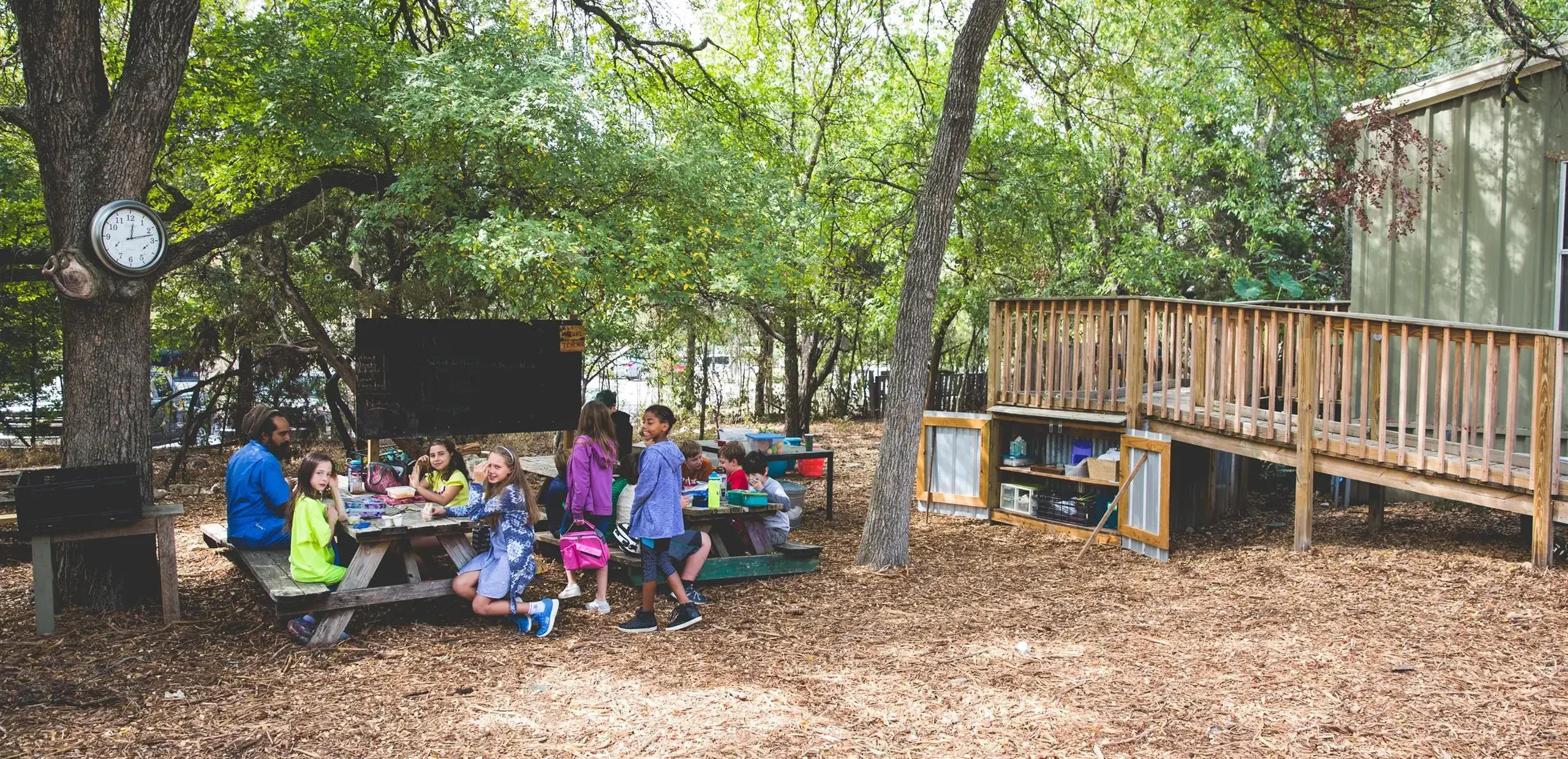 Students and a teacher gathered at picnic tables in a wooded outdoor classroom area near a chalkboard and elevated deck.