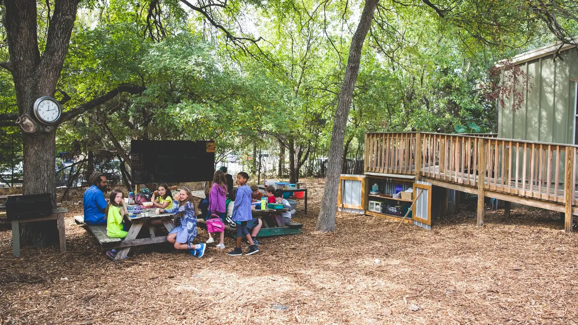 Students gathered at a picnic table in a wooded outdoor area near a deck.