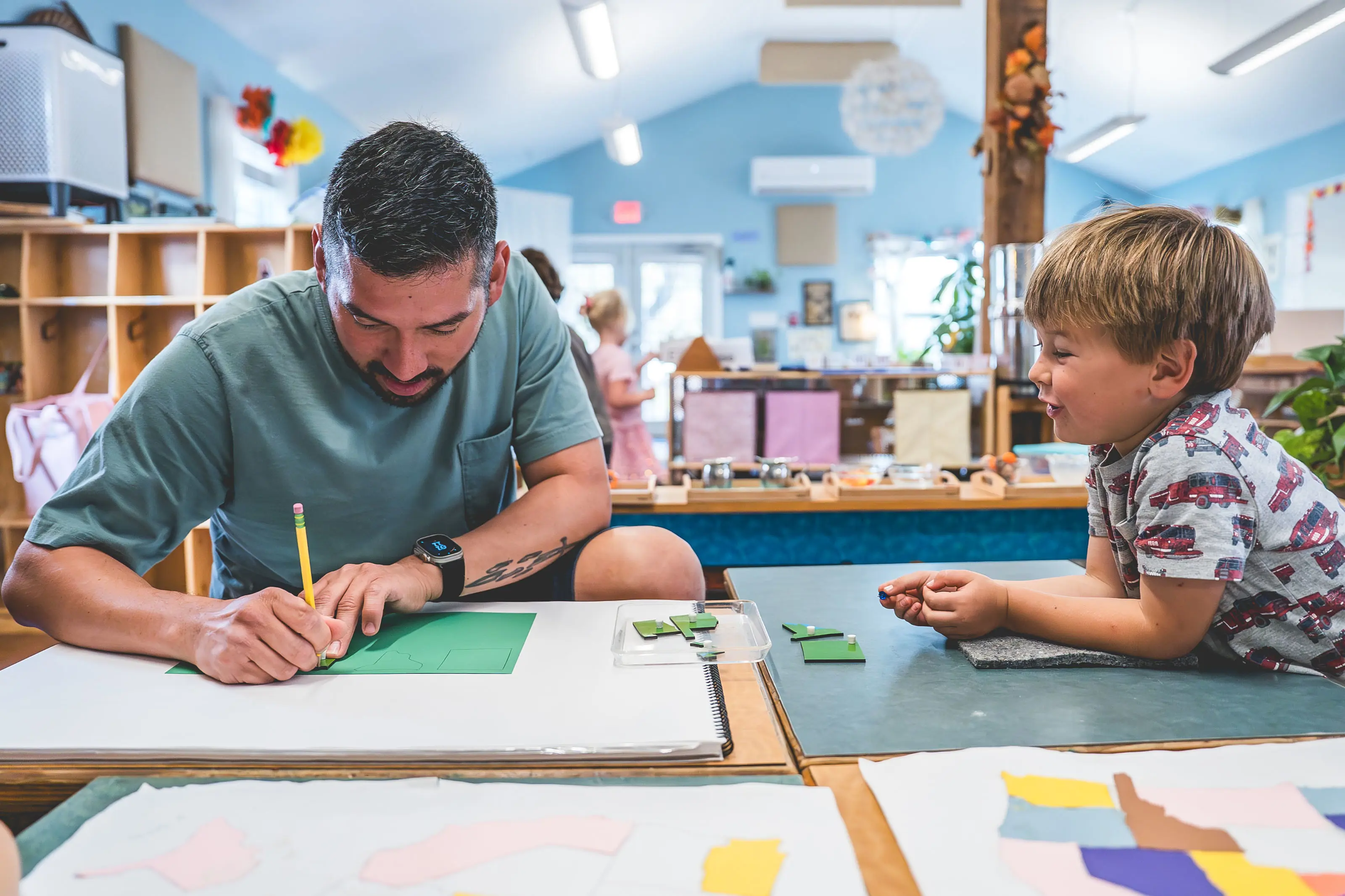 A teacher guiding a young student through a cutting activity at a table.