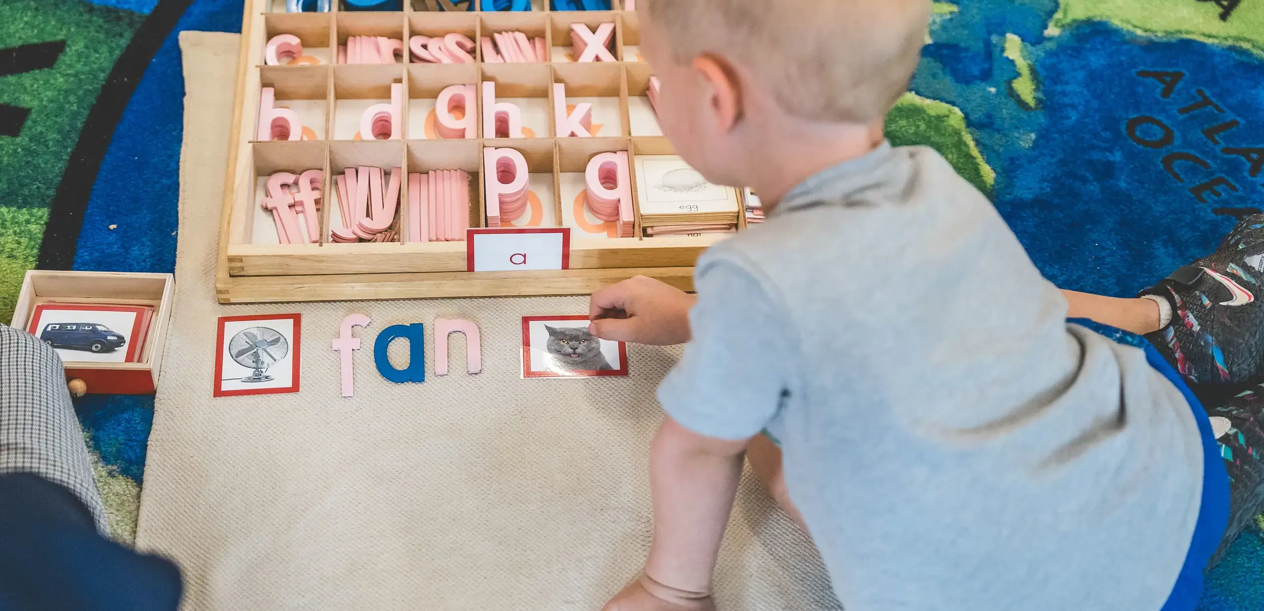 Young child using Montessori moveable alphabet letters to build the word 'fan' beside matching picture cards on a world map rug