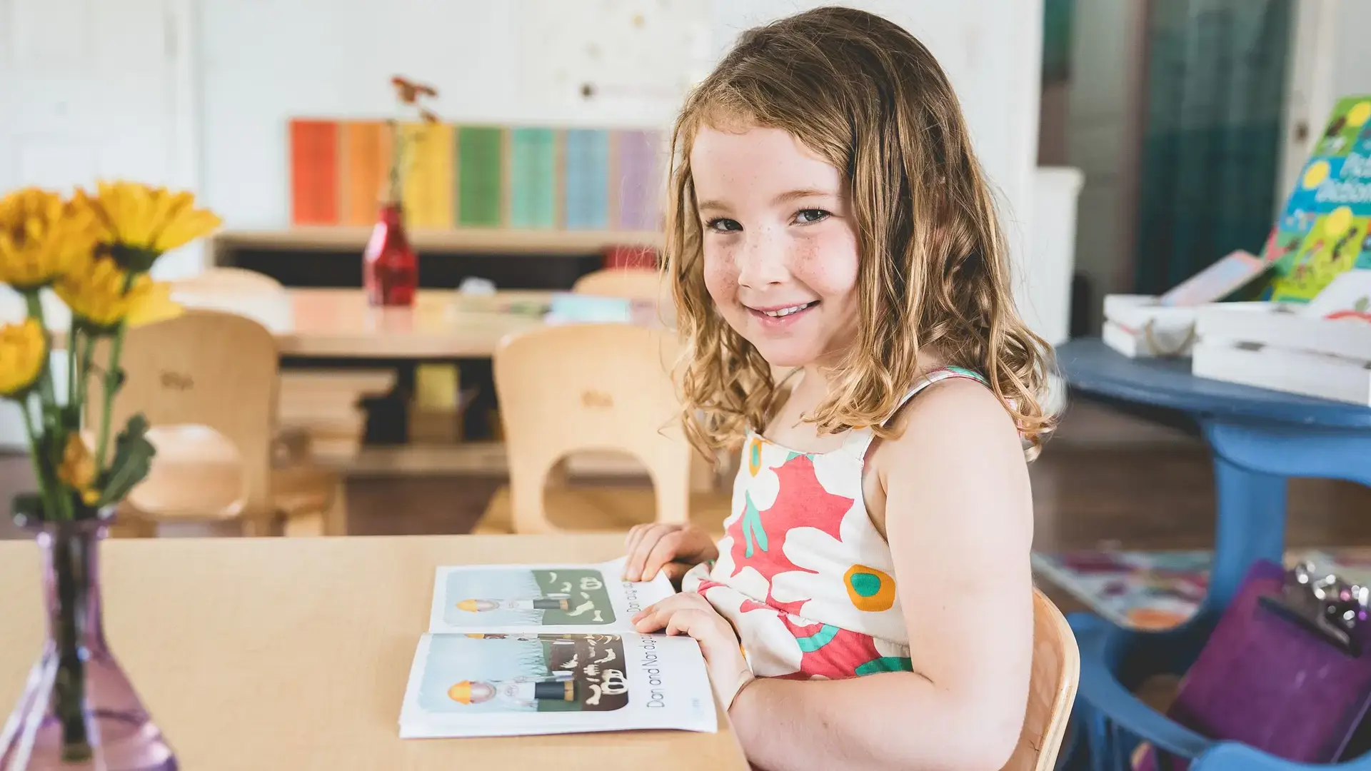 Young child smiling while reading a book at a table with flowers nearby.