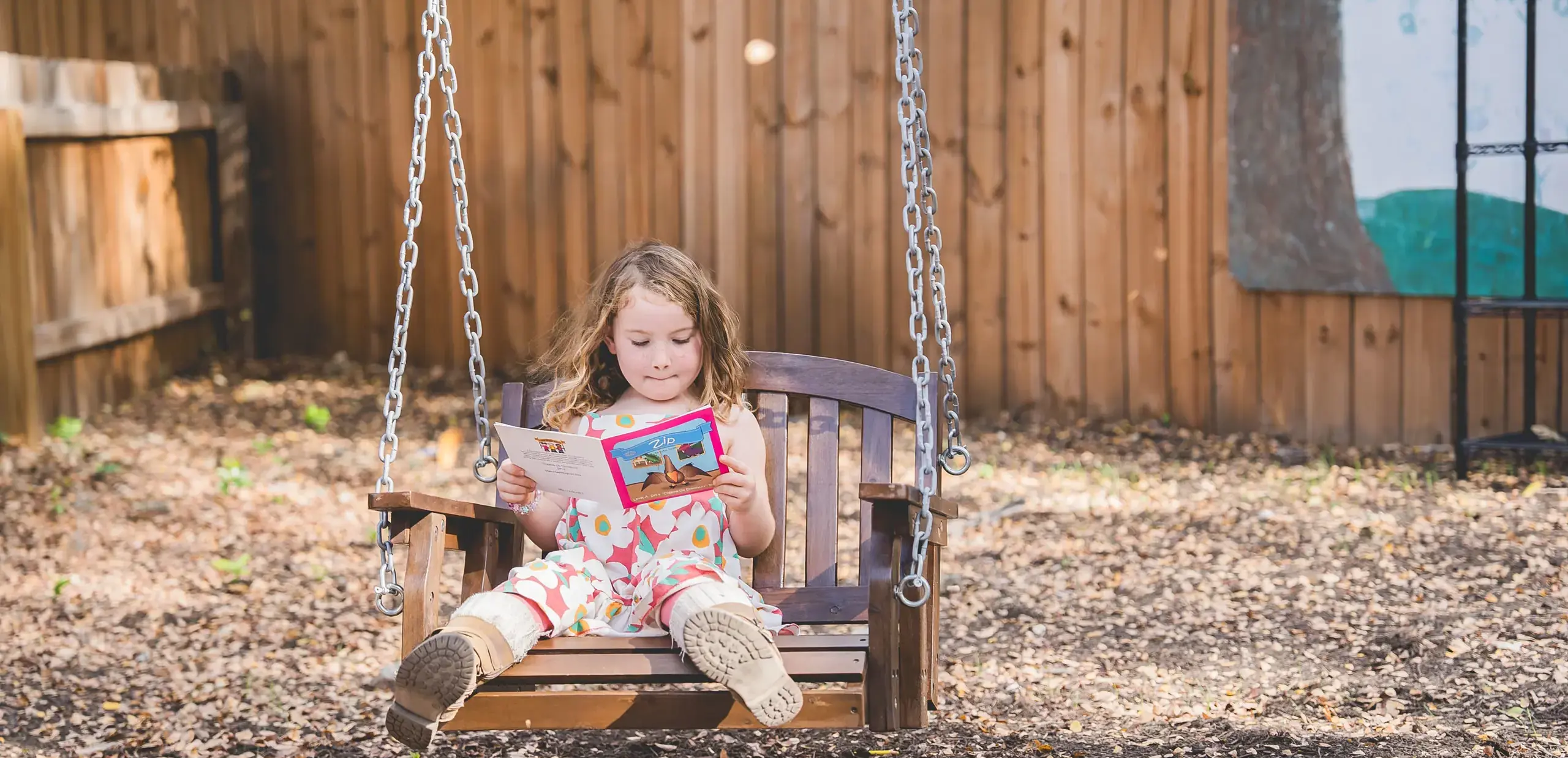 Young child reading a picture book while sitting on an outdoor wooden bench swing.