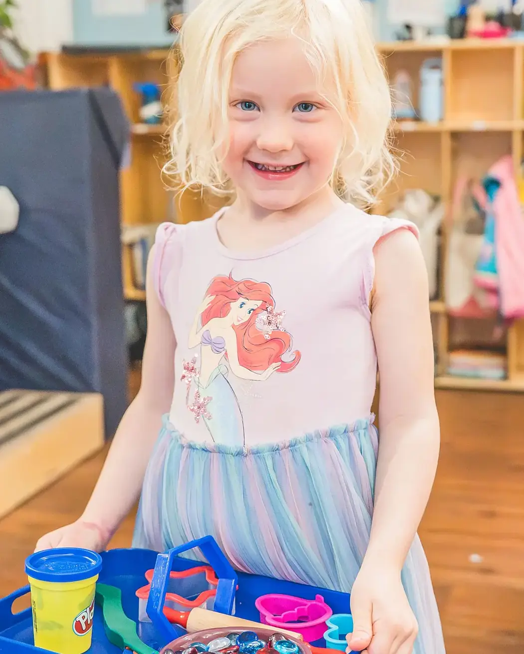 young student smiling while holding a tray of playdough and sensory tools in the classroom.