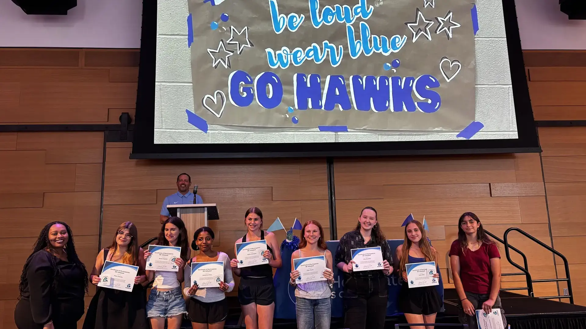 high school students holding certificates in front of a 'Go Hawks' banner during an athletics awards ceremony.