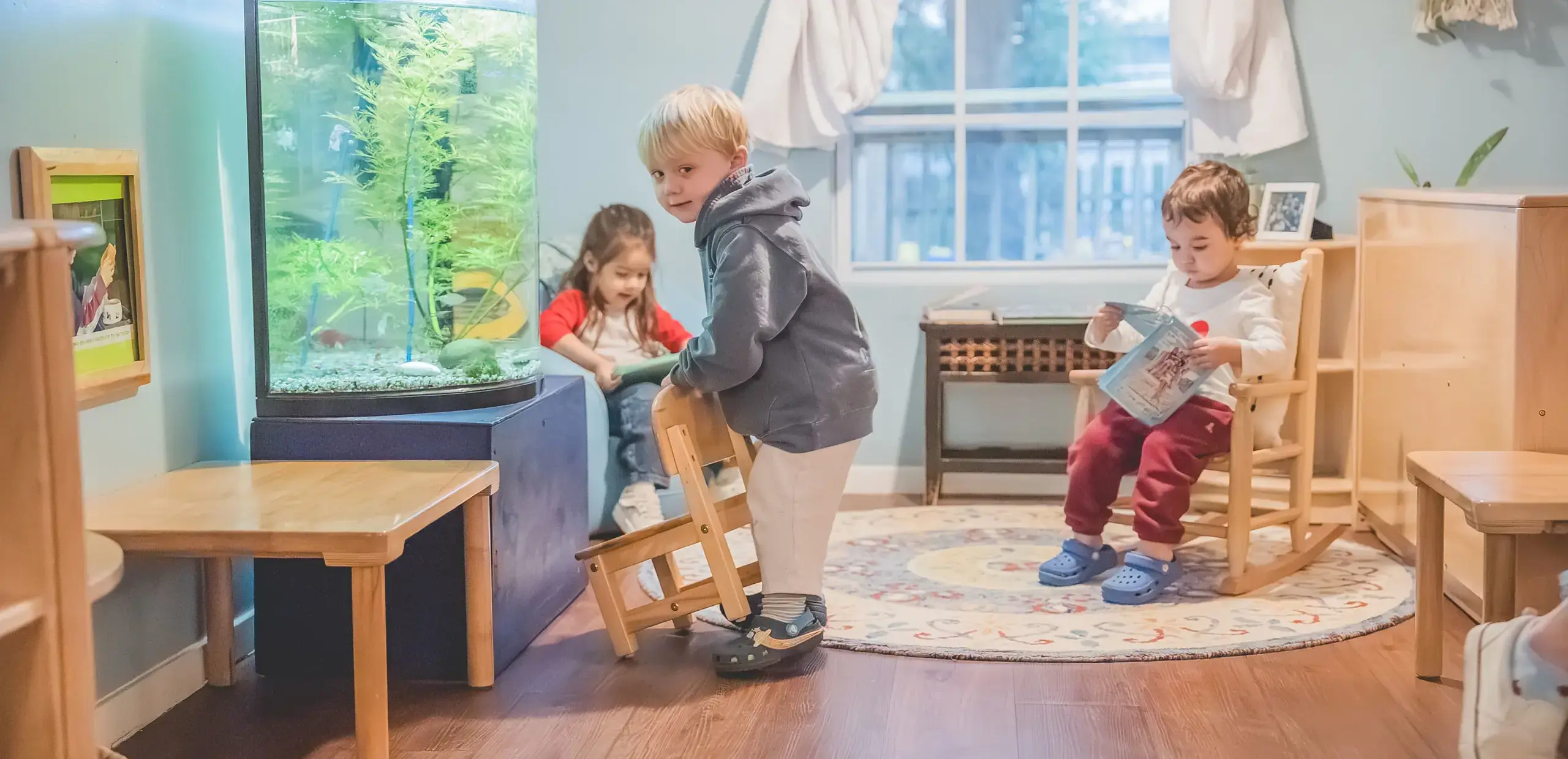 Young children in a classroom near a fish tank, with one child reading a book in a small rocking chair.