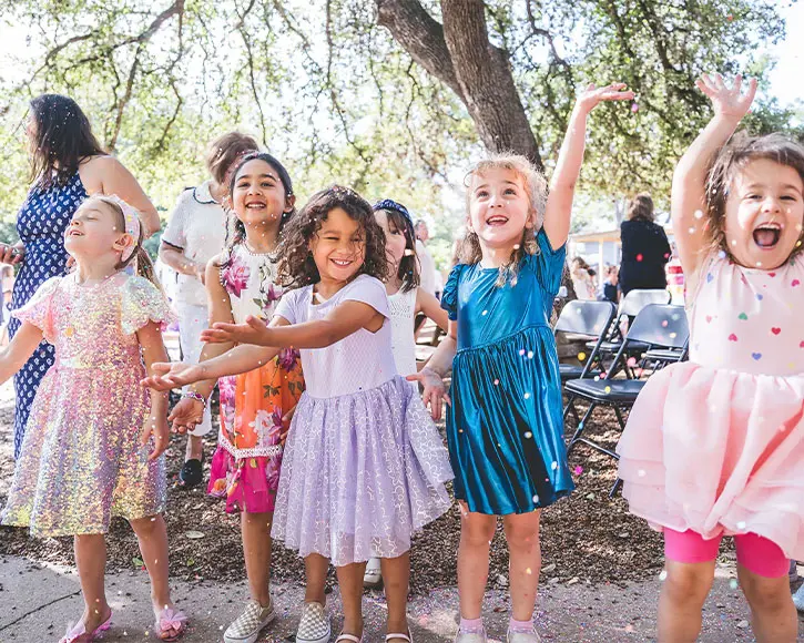 group of young girls in colorful party dresses standing outdoors under trees, excitedly reaching up and smiling as confetti falls around them, with adults and folding chairs visible in the background.